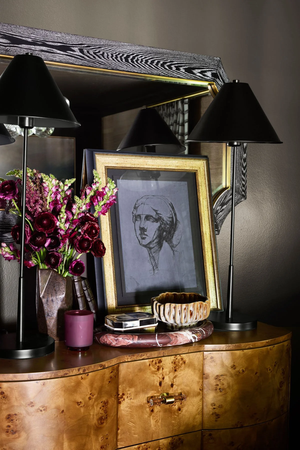 A wooden dresser with a granite top holds a framed sketch of a woman's face, black table lamps, a vase of purple and pink flowers, a pink candle, and ceramic bowls, with a large mirror above reflecting the room.