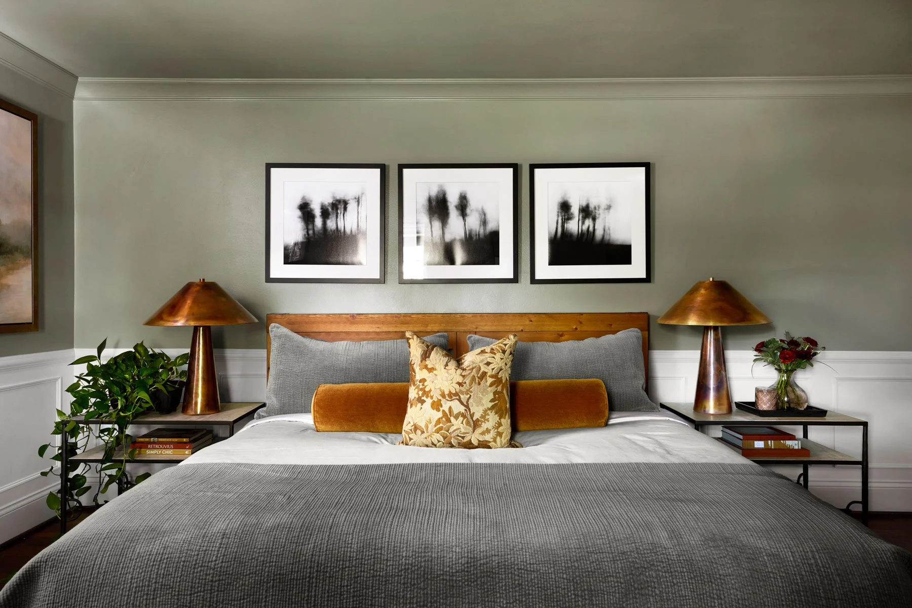 A neatly made bed with gray and orange pillows, flanked by wooden nightstands with brass lamps, framed black-and-white abstract art above, greenery and a vase with red roses on the right, and books on the left.