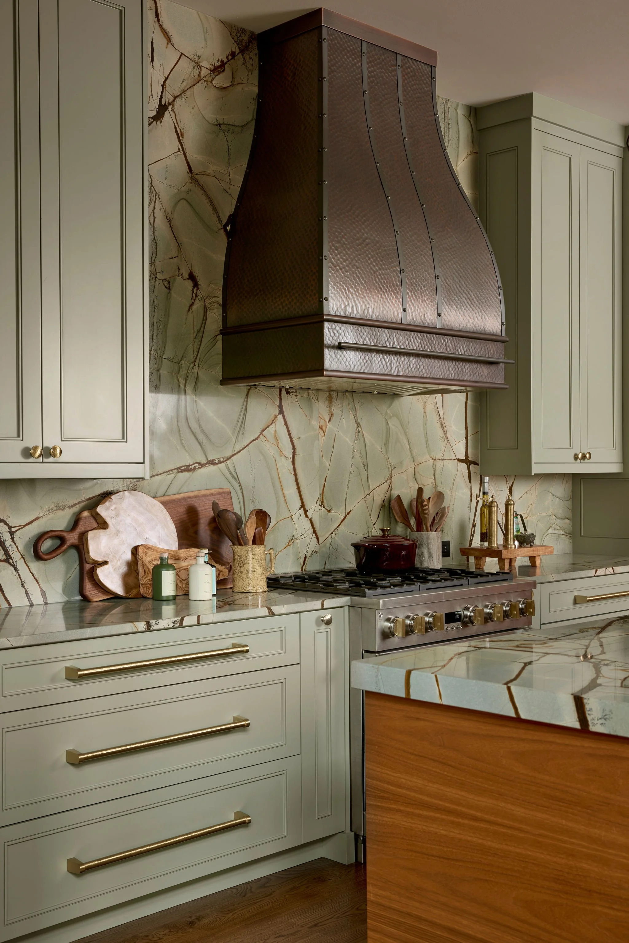 A kitchen with cream-colored cabinets, a marble countertop, and a marble backsplash with brown and beige veining. There is a large copper range hood above a stainless steel stove, and decorative kitchen items such as cutting boards, utensils, and bottles are on the counter.