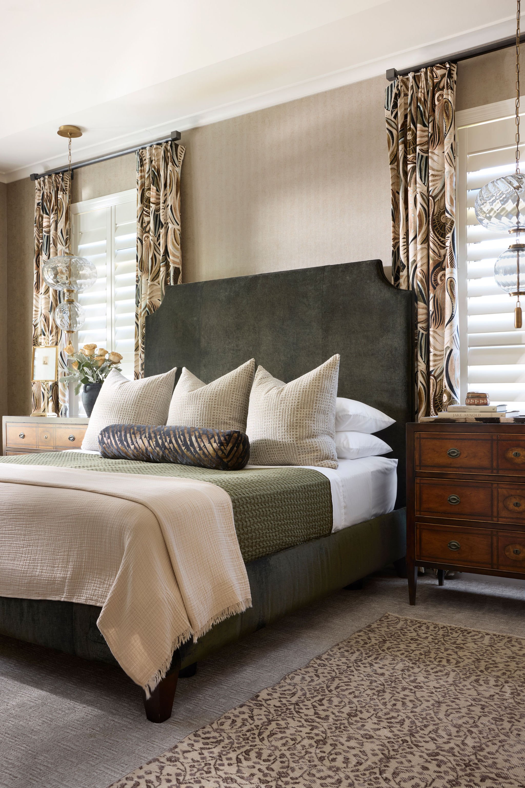 A bedroom with a large dark upholstered bed, beige decorative pillows, and a beige throw blanket. There are patterned curtains, a wooden nightstand with books and a table lamp, and a window with white plantation shutters.