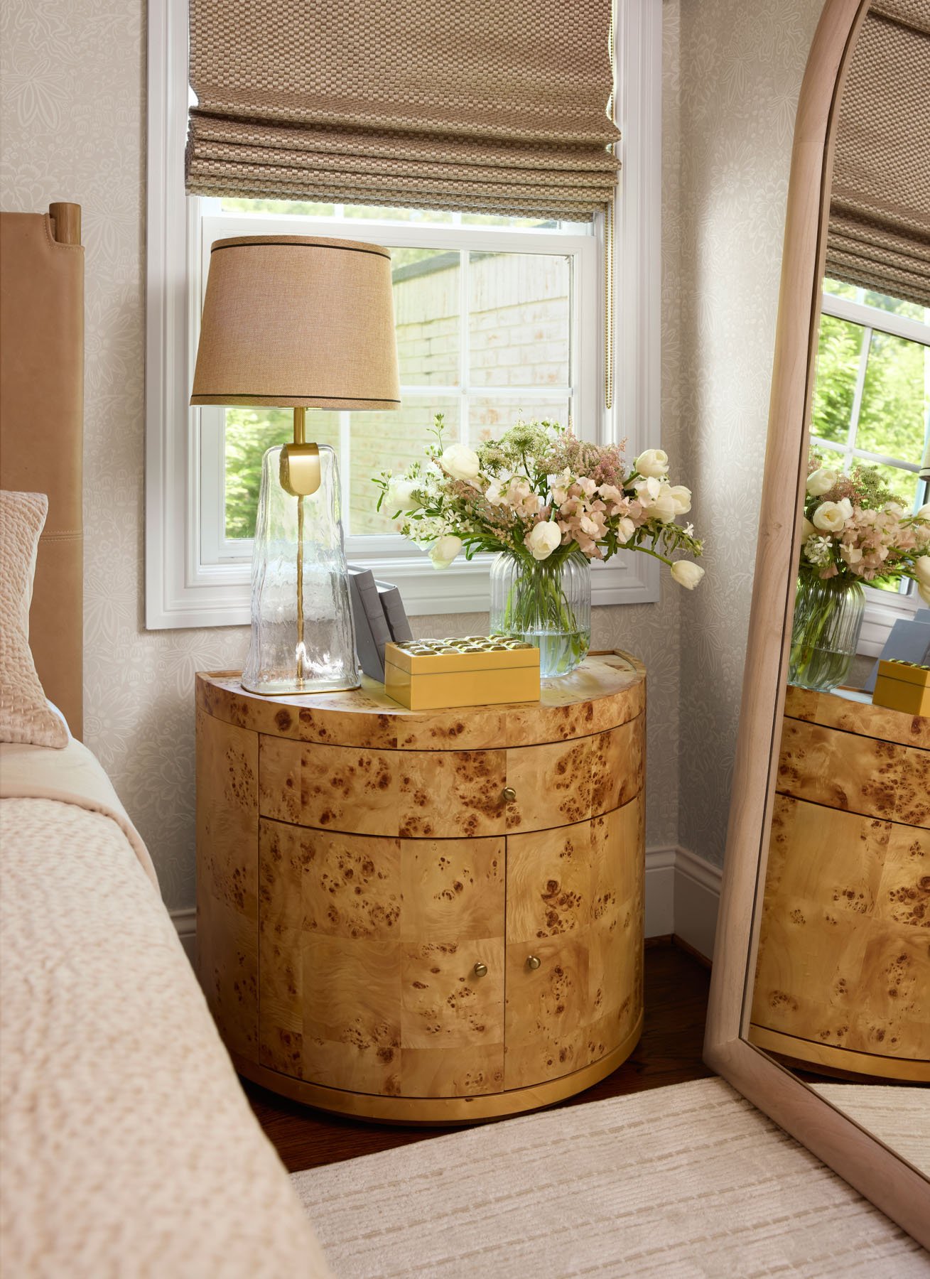 A bedroom side table made of light wood with a curved front, decorated with a beige table lamp, a glass vase with white and pink flowers, and a small yellow box. There is a window with beige Roman shades behind, and a mirror reflects part of the room