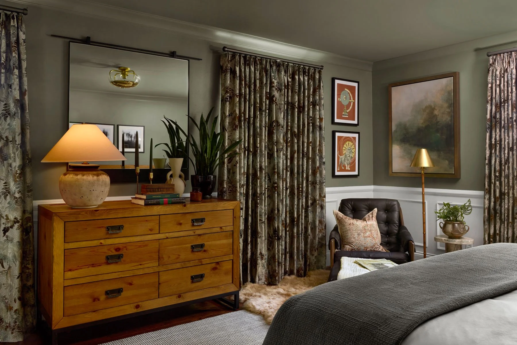 A cozy bedroom corner with a wooden dresser, decorative objects, a mirror, a tall plant, framed artwork, a brown leather armchair with a patterned pillow, a floor lamp, a small table with a potted plant, and floral curtains.