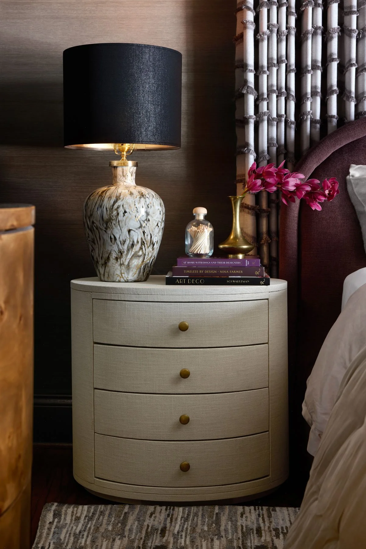 A bedroom nightstand with a marble lamp, a stack of books, a small glass container of toothpicks, and a gold vase with pink flowers, beside a bed and a curtain.