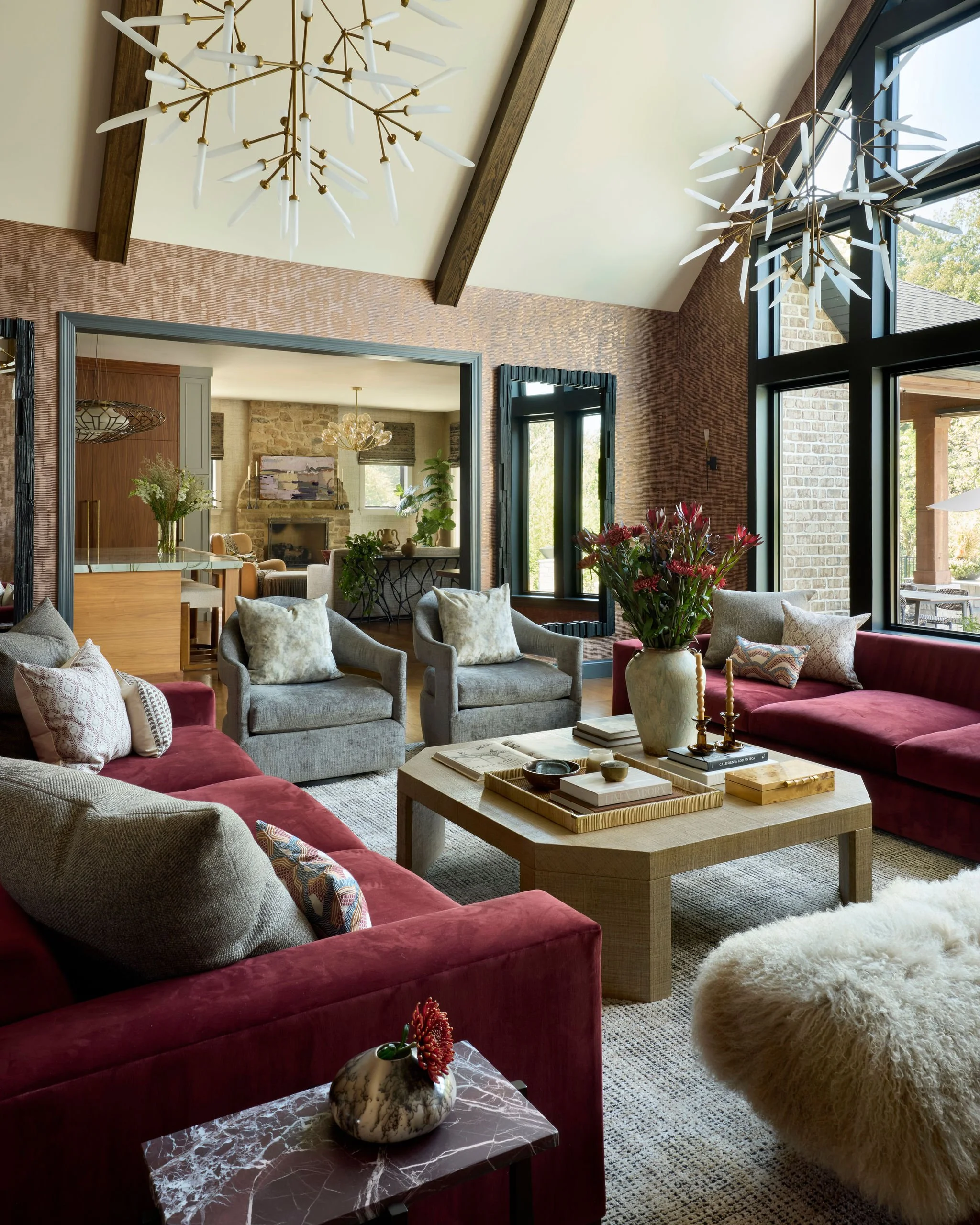 A living room with red velvet sofas, gray armchairs, a wooden coffee table with decorative items, large windows, and modern chandeliers.