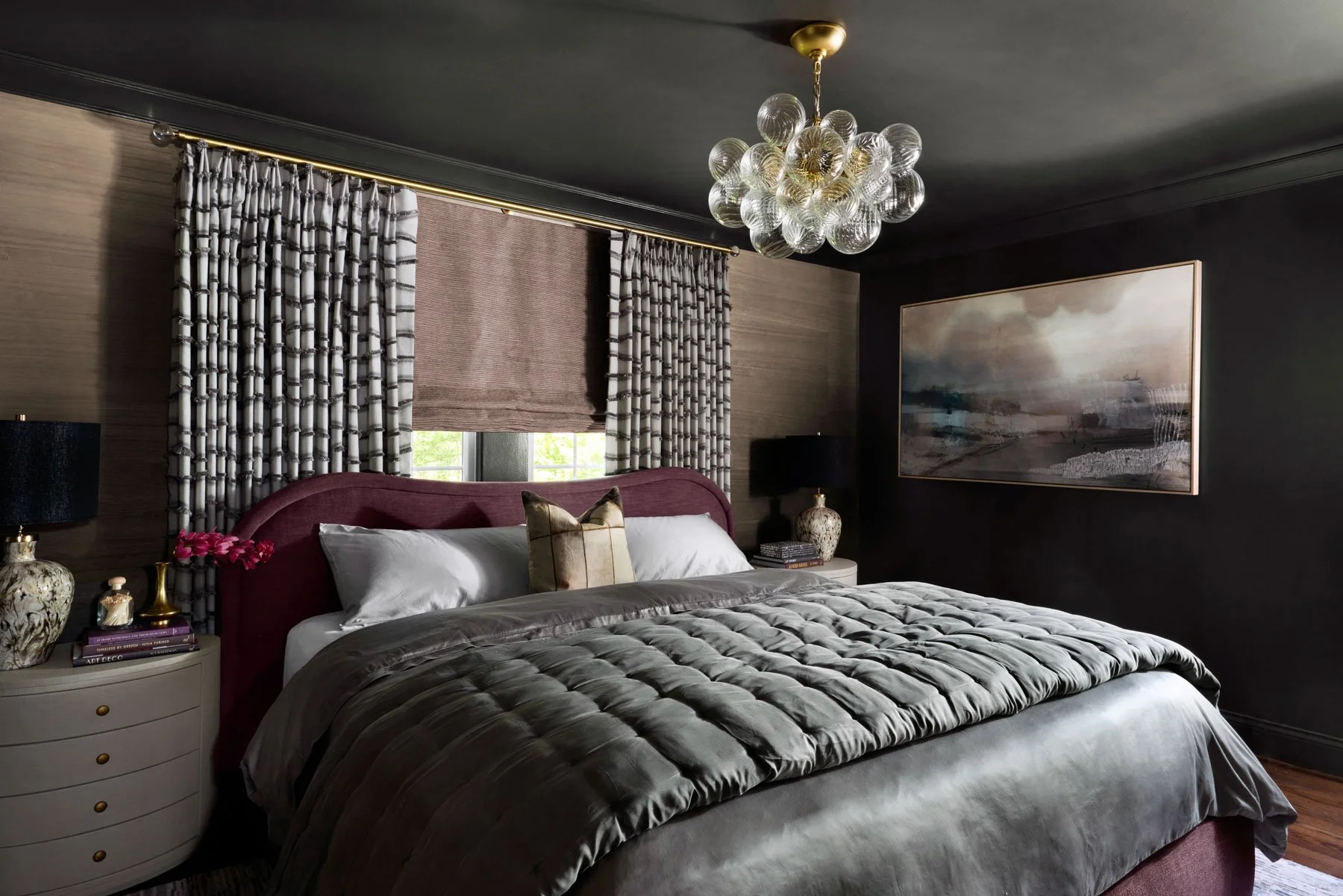 A bedroom with a burgundy upholstered headboard, white bedding, and a gray velvet comforter. Two black lamps, decorative vases, and books are on the bedside tables. There are patterned curtains and a window, with a large abstract painting on the dark