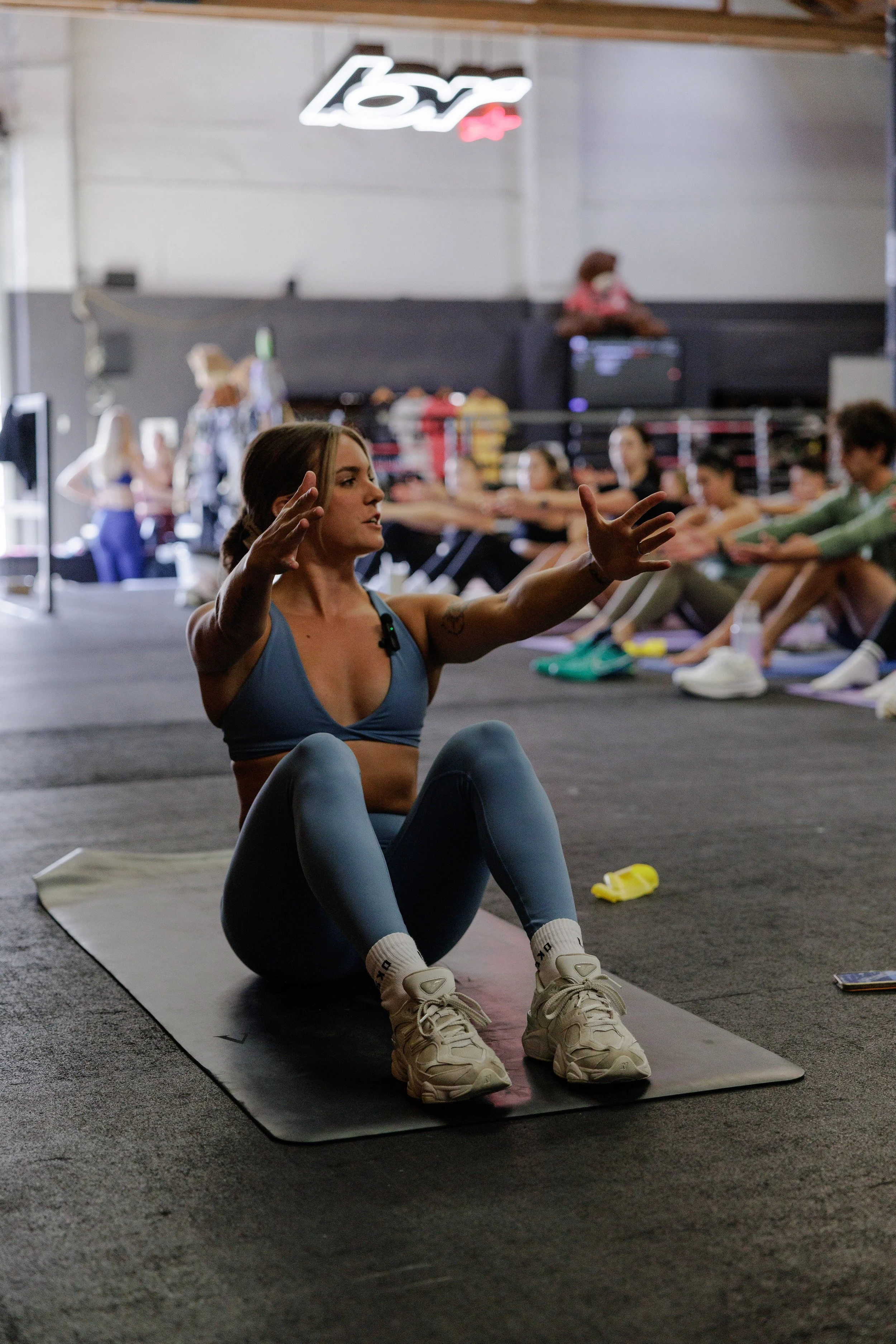 A woman exercising on a mat in a gym, hosting a large amount of people.