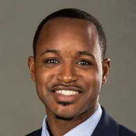 Close-up of a smiling African American man in a suit against a plain gray background.