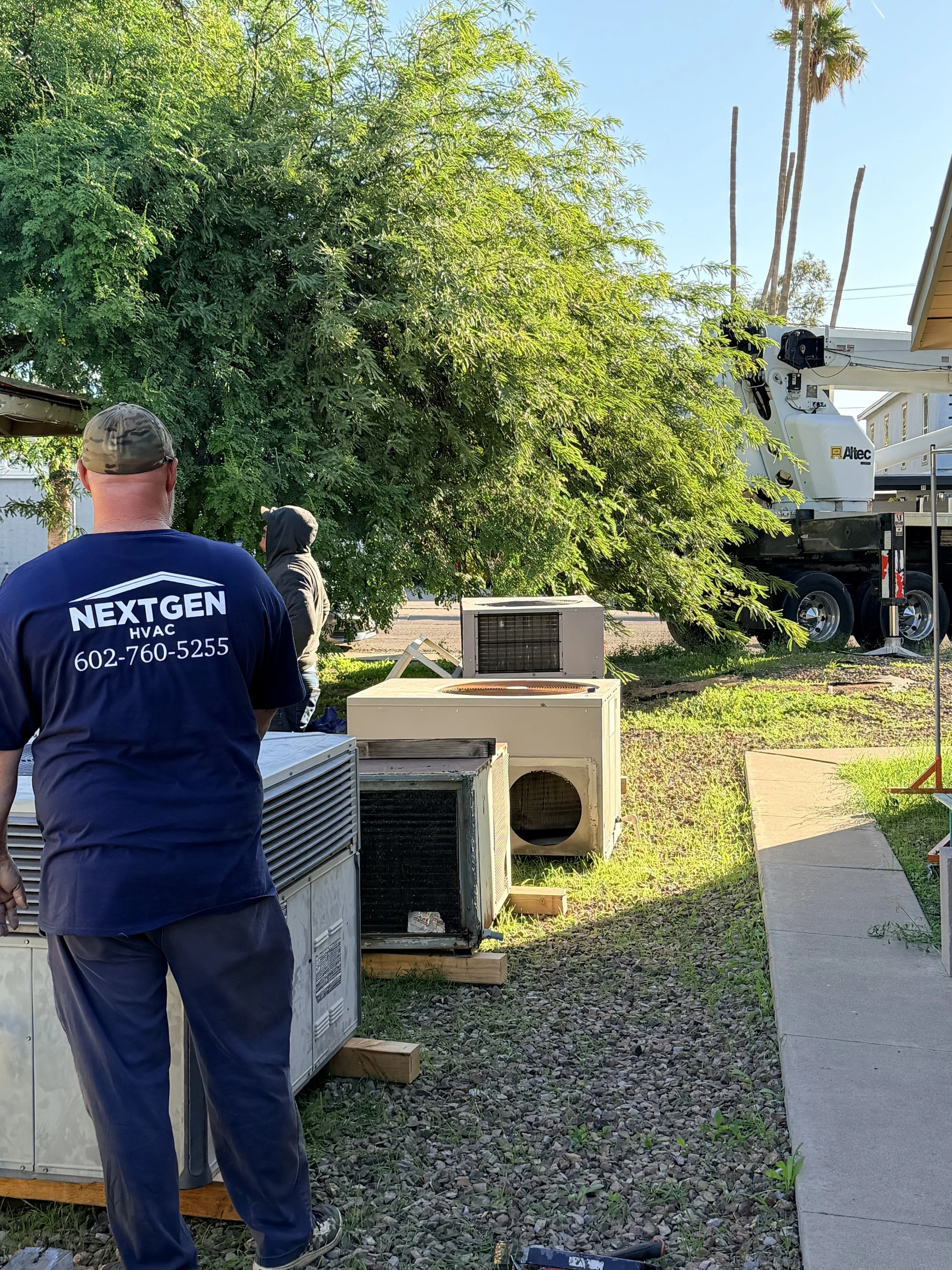 Technicians working outdoors next to discarded air conditioning units, with a large tree and a crane truck in the background.