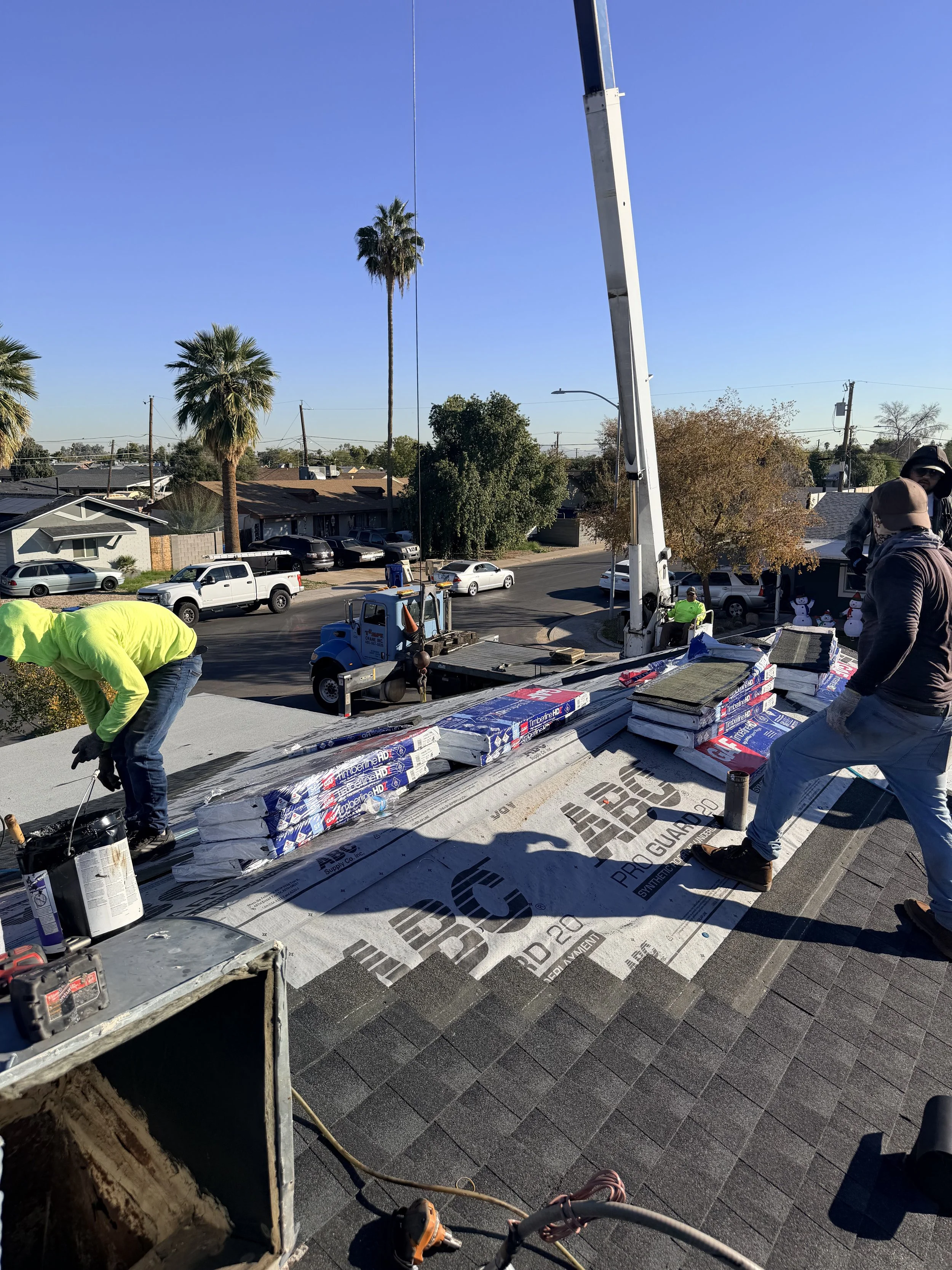 Roofing workers installing shingles on a house roof with a crane and construction materials around.