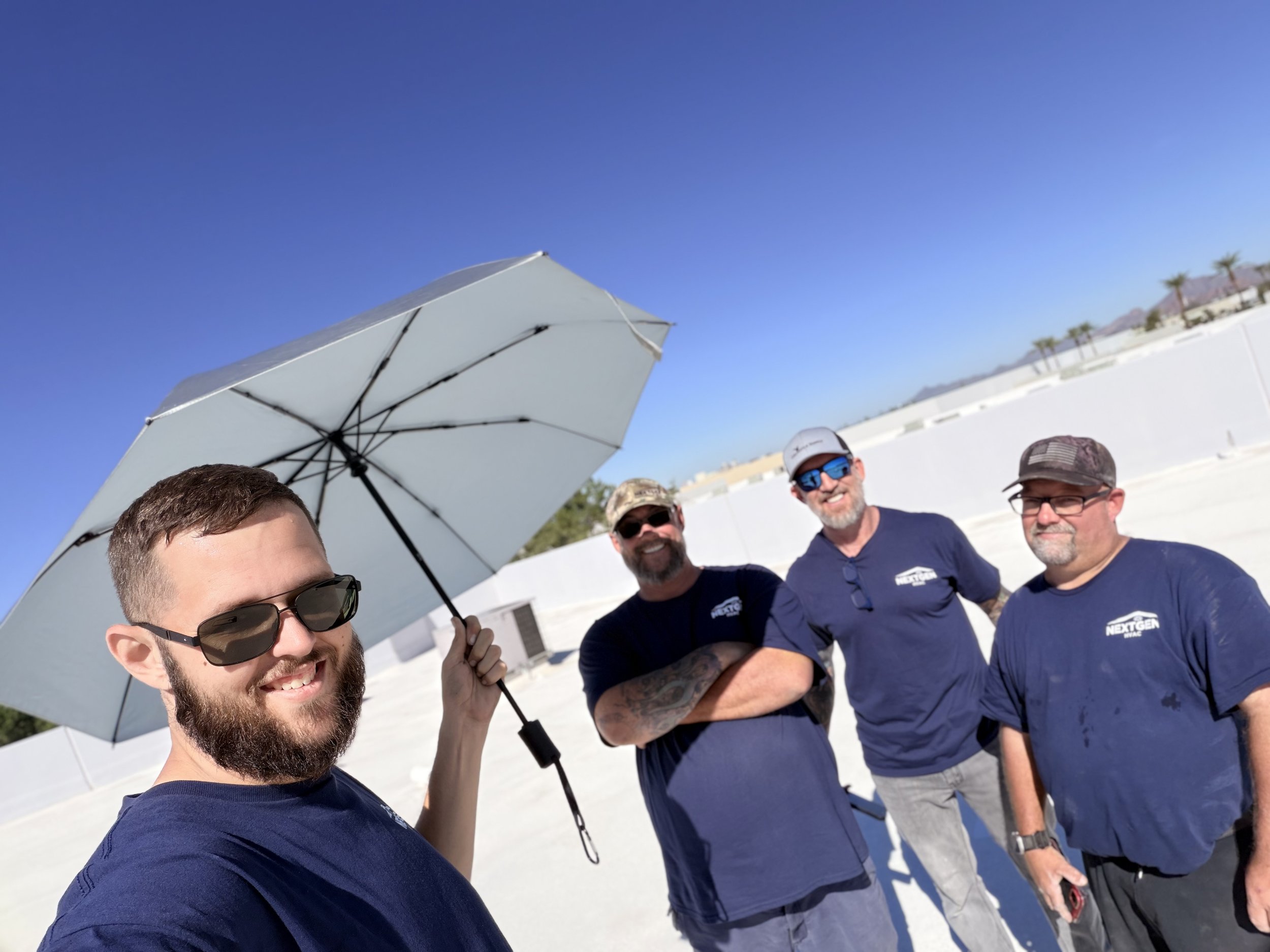 Four smiling men standing outdoors on a sunny day, with one holding an umbrella, all wearing casual clothes and sunglasses.