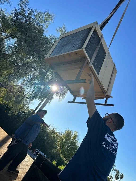Man lifting a small wooden house or cabin with a crane outdoors on a sunny day, with trees and a clear blue sky in the background.