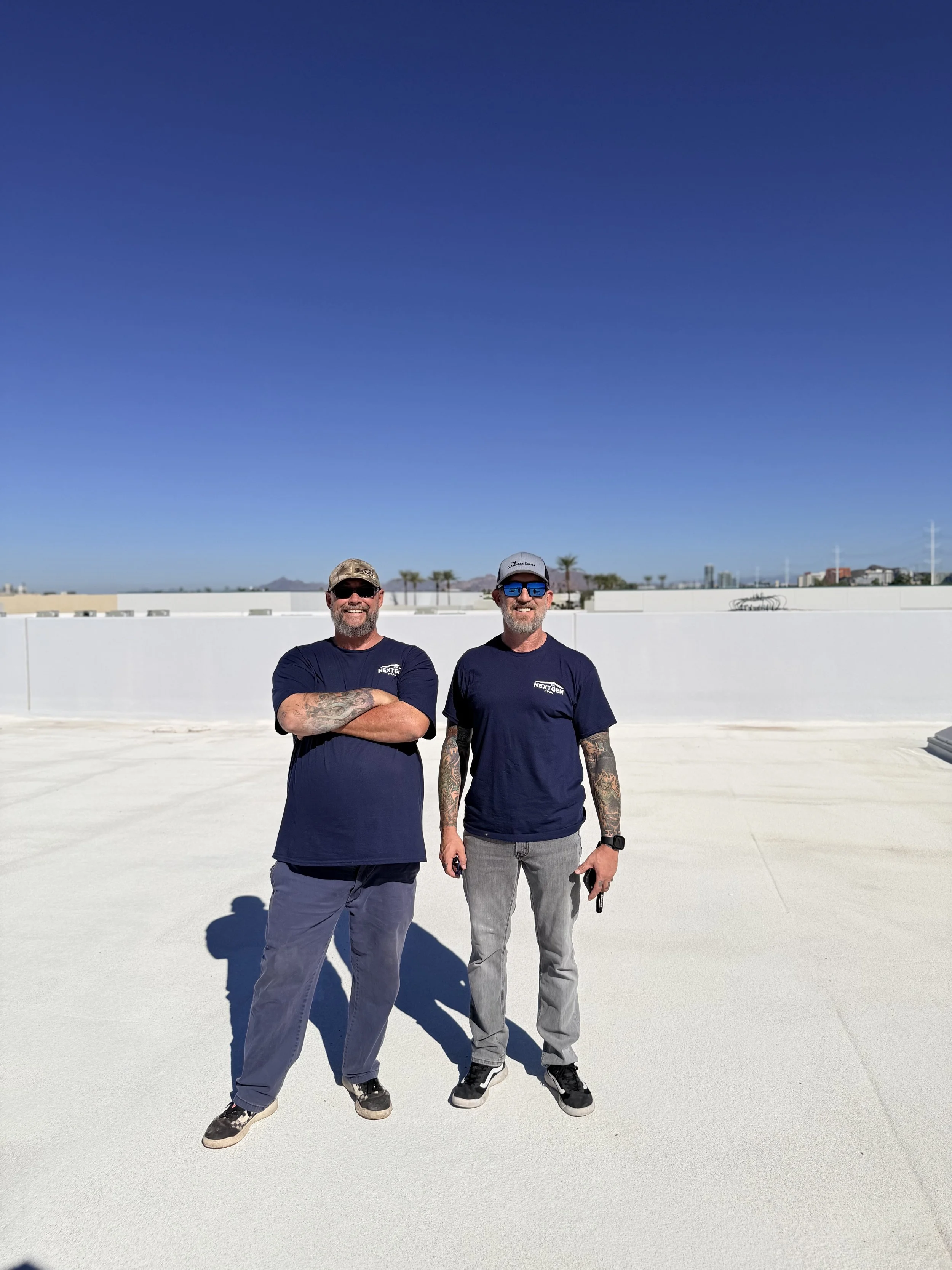 Two men standing on a rooftop, smiling, with a clear blue sky and palm trees in the background. They wear navy t-shirts and sunglasses.