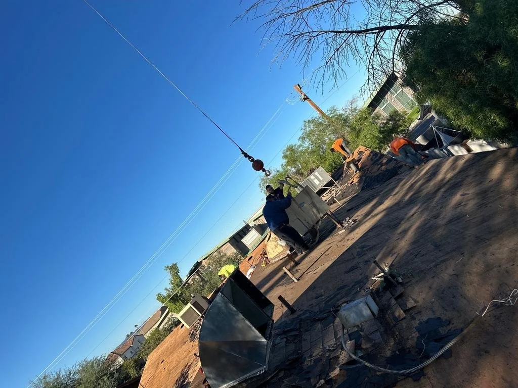 Construction workers install utility poles on a dirt lot under a clear blue sky, with trees and nearby rooftops in the background.