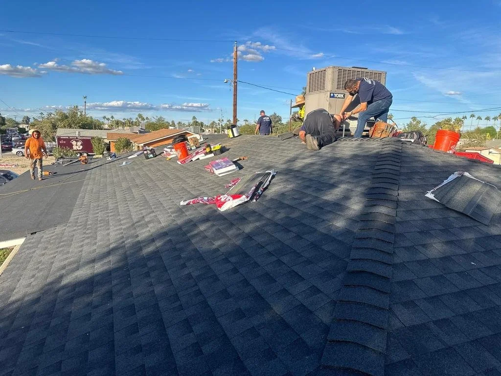 Workers installing or repairing a dark asphalt shingle roof on a building, with roofing tools and materials scattered around, under a clear blue sky.