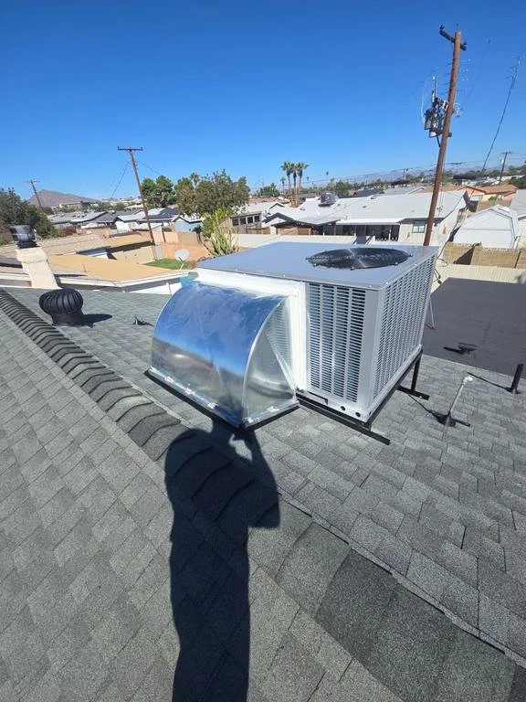 View of a rooftop with HVAC units, including a large air conditioning unit and a smaller, curved vent, under a clear blue sky in a residential neighborhood.