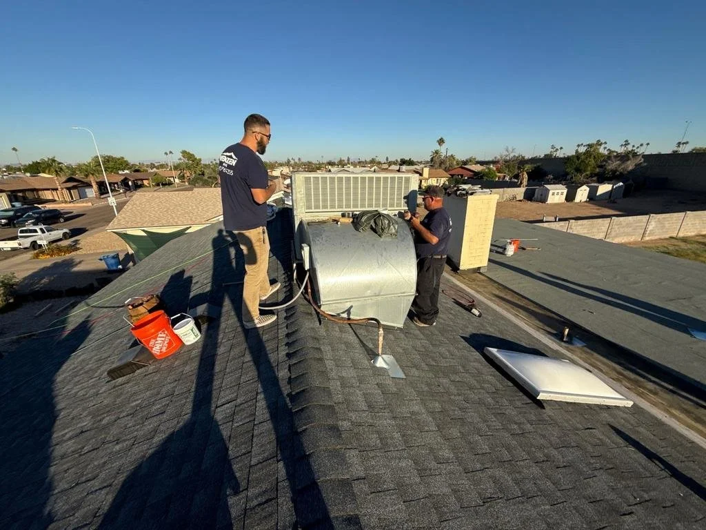 Two workers installing or repairing a ventilation unit on a residential roof during daytime. One worker is standing on the roof, the other is on the ground, surrounded by tools and equipment.