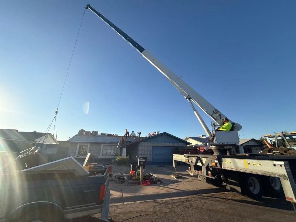 A crane lifting a boat in a residential neighborhood with workers on the roof and a truck parked on the street.