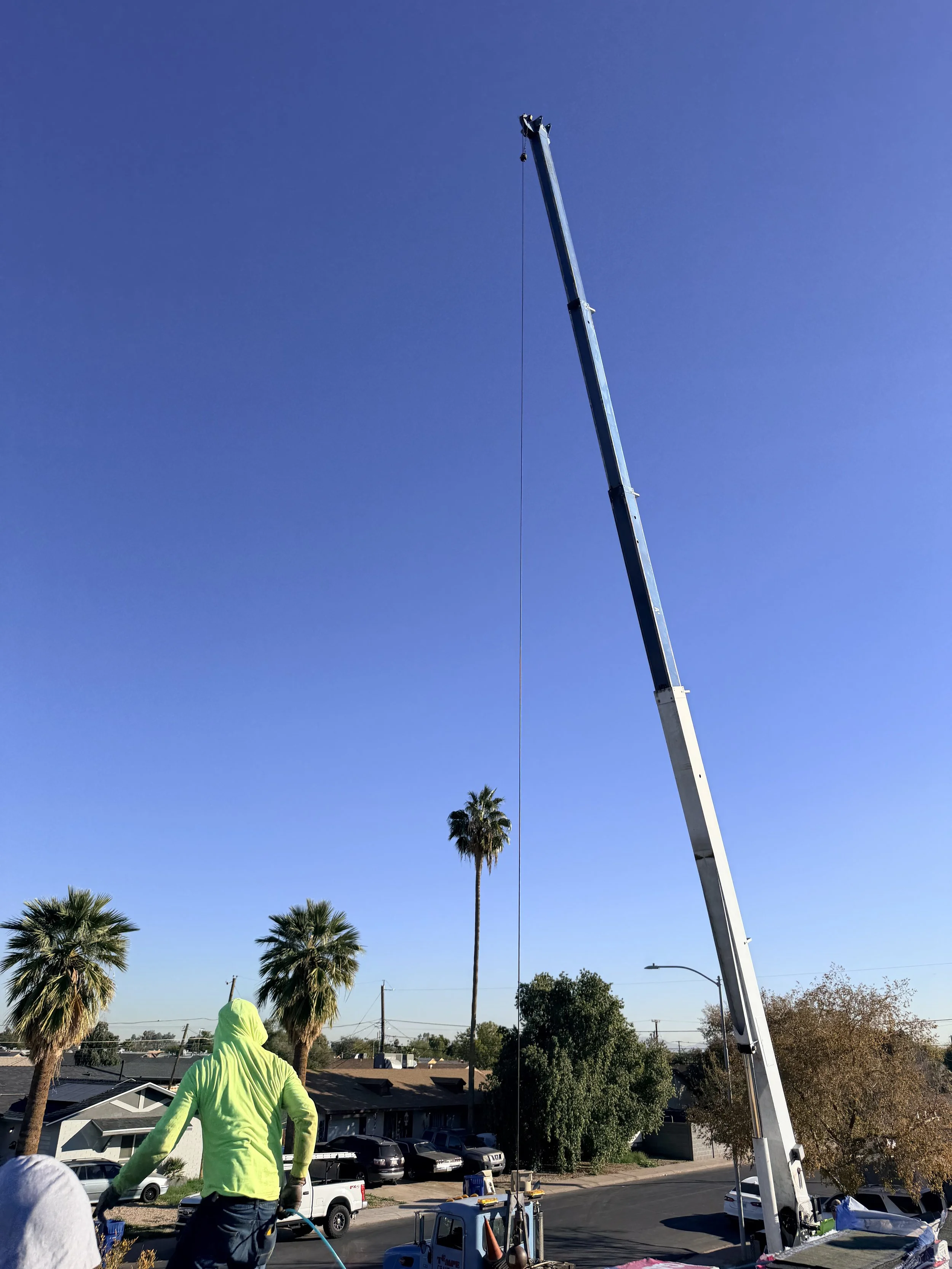 Worker in bright green jacket operating a crane on a residential street with palm trees against a clear blue sky.