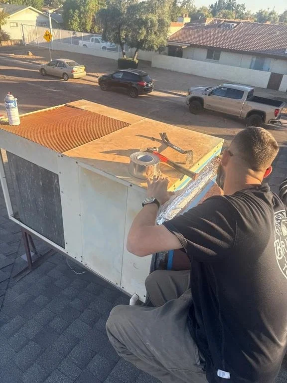 A man is working on the roof of a small building or kiosk, installing a reflective foil material with tools placed on top, overlooking a street with parked cars and trees in the background.