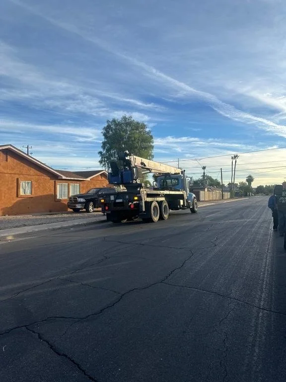 A large utility crane truck parked on a city street with residential houses and trees in the background under a blue sky with wispy clouds.