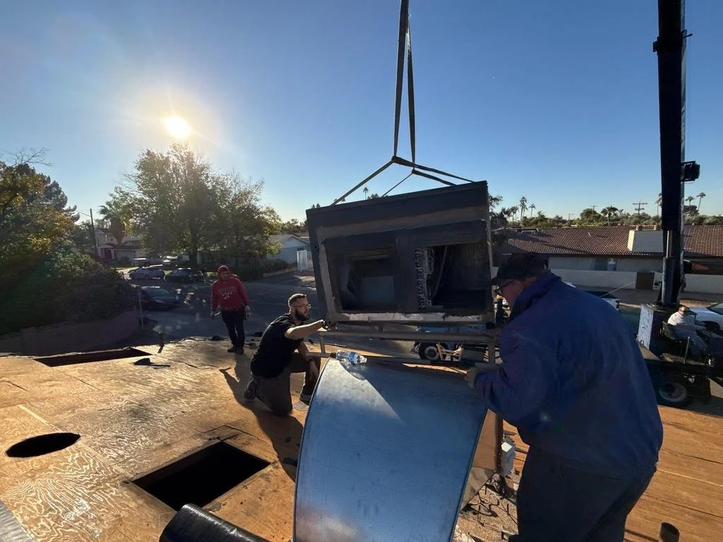 Workers install a large air conditioning unit on a rooftop during daylight, with a few other people and parked cars visible in the background.