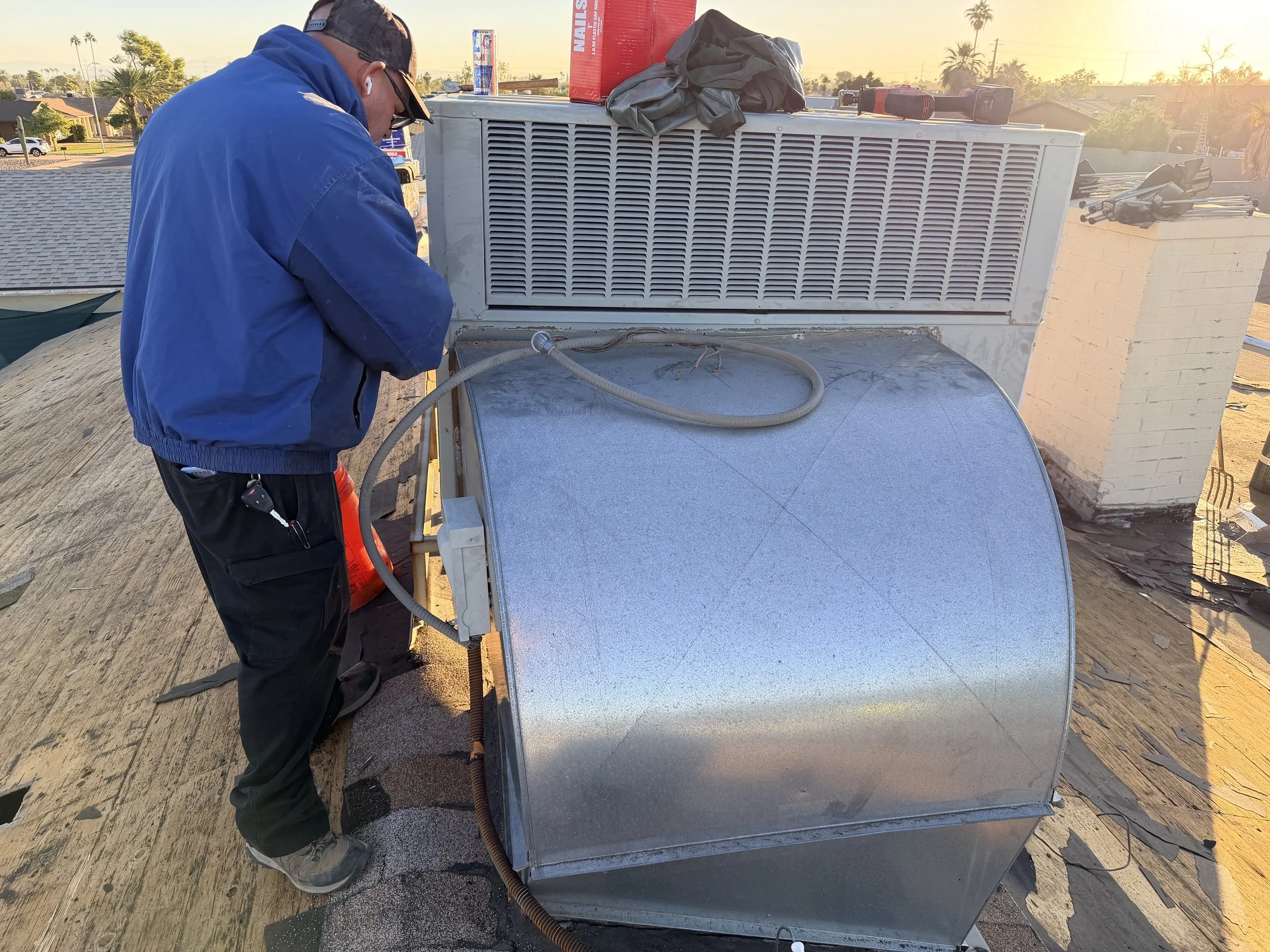 A technician working on an air conditioning unit on a rooftop during sunset, wearing a blue jacket and black pants.