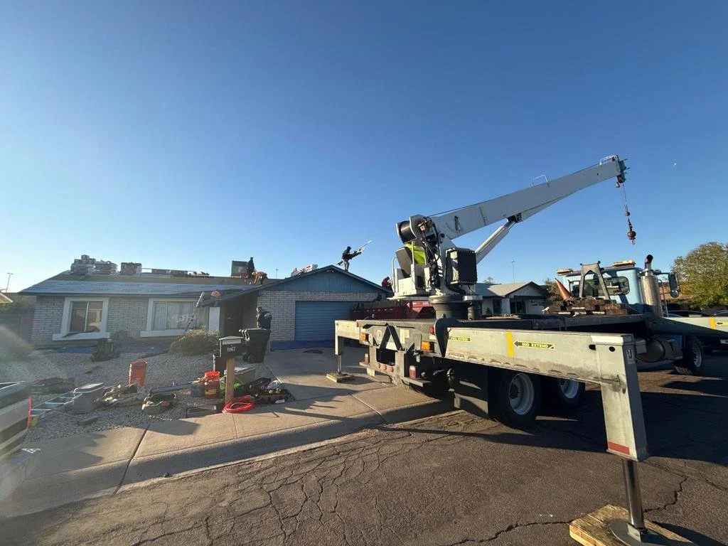 A house with a crane on a truck lifting items through the roof during roof repair or replacement, with workers on the roof and tools scattered on the driveway.