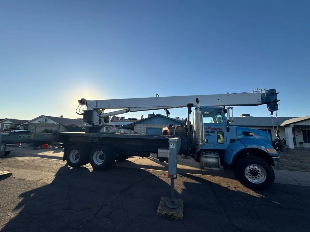 A blue crane truck parked on a residential street with houses in the background, the sun setting behind the truck.