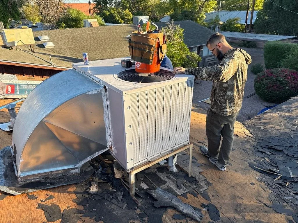 Two workers are repairing or installing HVAC equipment on a rooftop, with one working on the unit and the other nearby. The roof has damaged shingles around the HVAC unit.