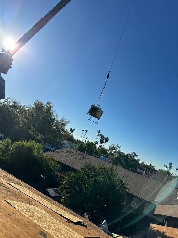 A construction crane lifting a small portable building in a suburban neighborhood with houses, trees, and clear blue sky.