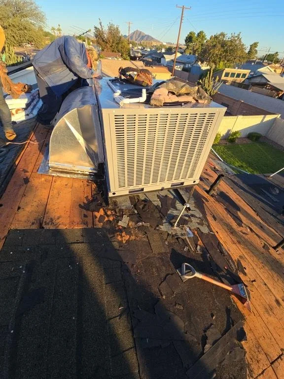 Workers repairing or replacing an HVAC system on a rooftop during daylight, with a clear sky and neighboring residential houses visible in the background.