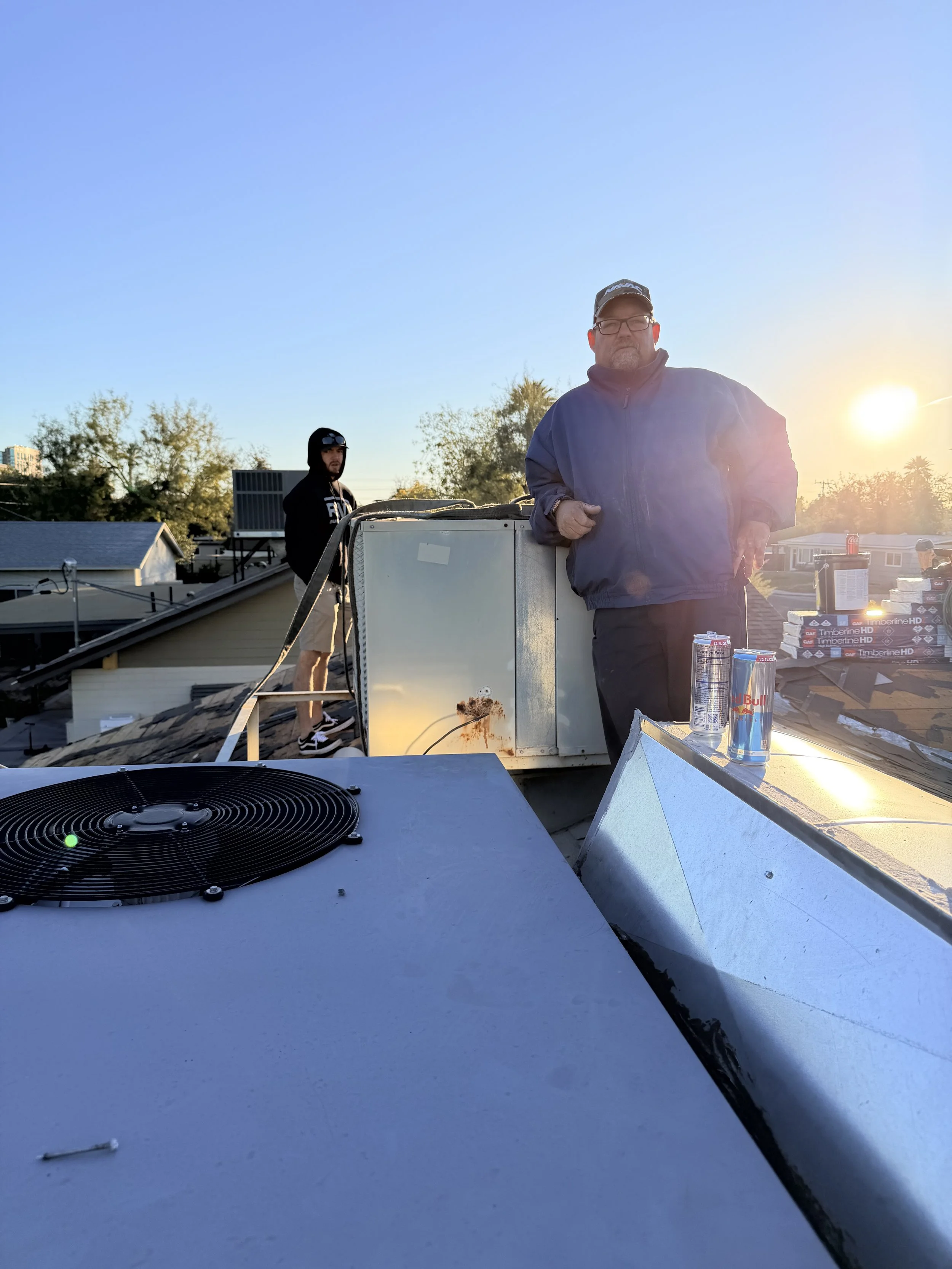 Two men on a rooftop during sunset, one in the foreground with glasses, dark jacket, and a cap, holding a tool, and the other in the background wearing a hoodie and shorts, near an air conditioning unit and cans of Red Bull, with boxes of pizza in the background.