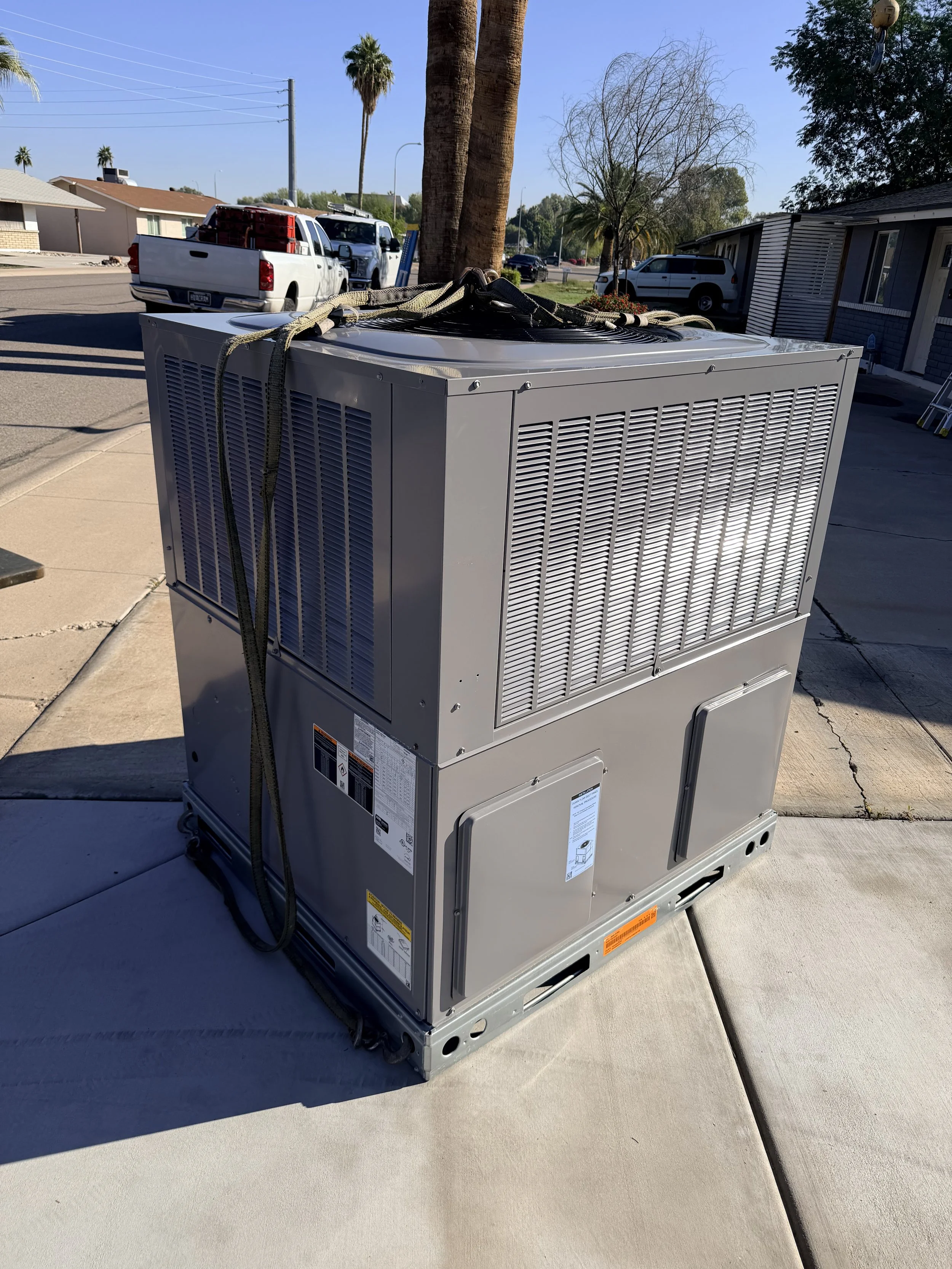 Large gray air conditioning unit installed on a concrete sidewalk outside a building, with a neighborhood street, cars, and palm trees in the background.