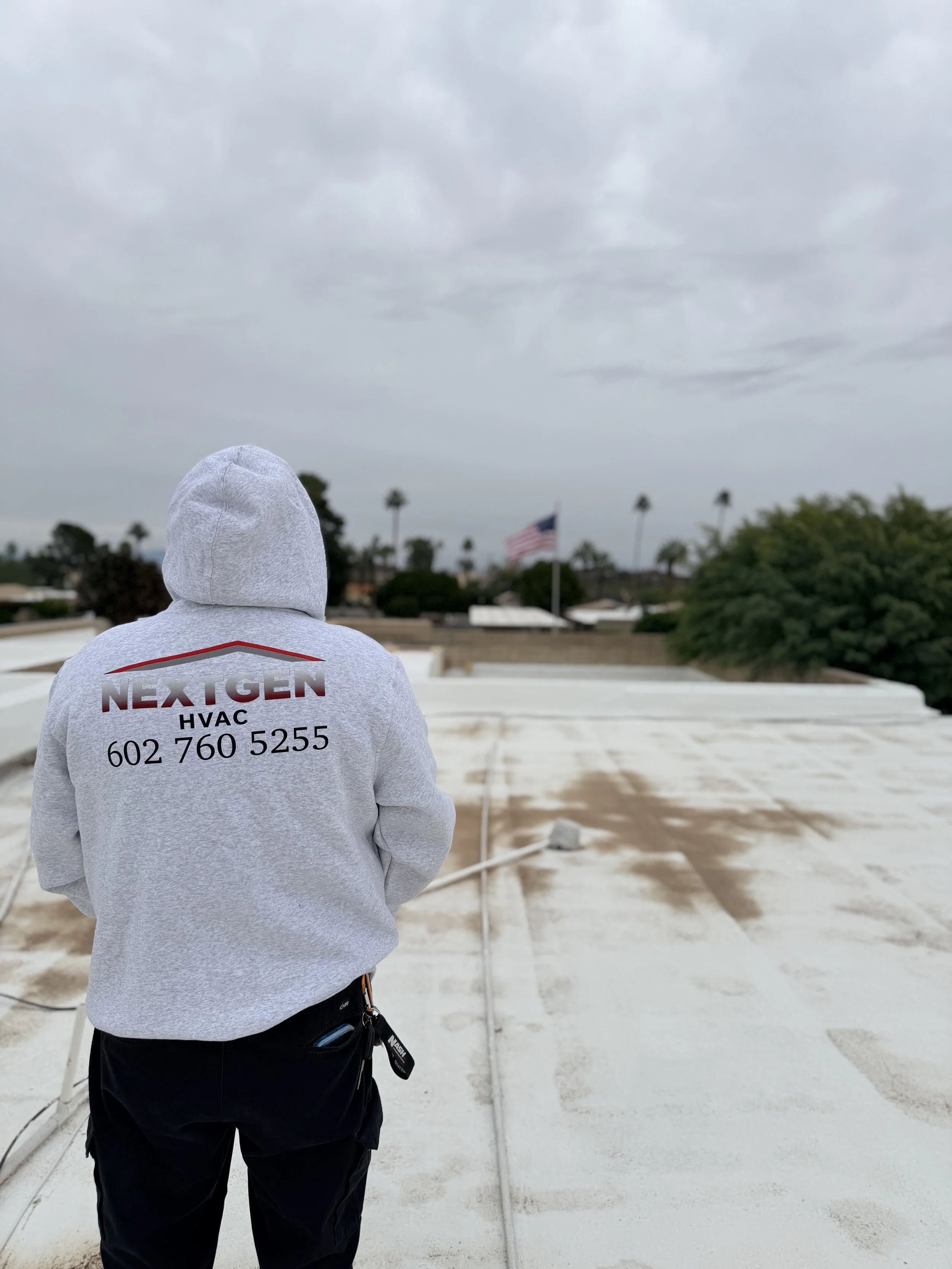 Person wearing a light gray hoodie with a logo and contact info on the back, standing on a rooftop under a cloudy sky, with palm trees and an American flag in the background.
