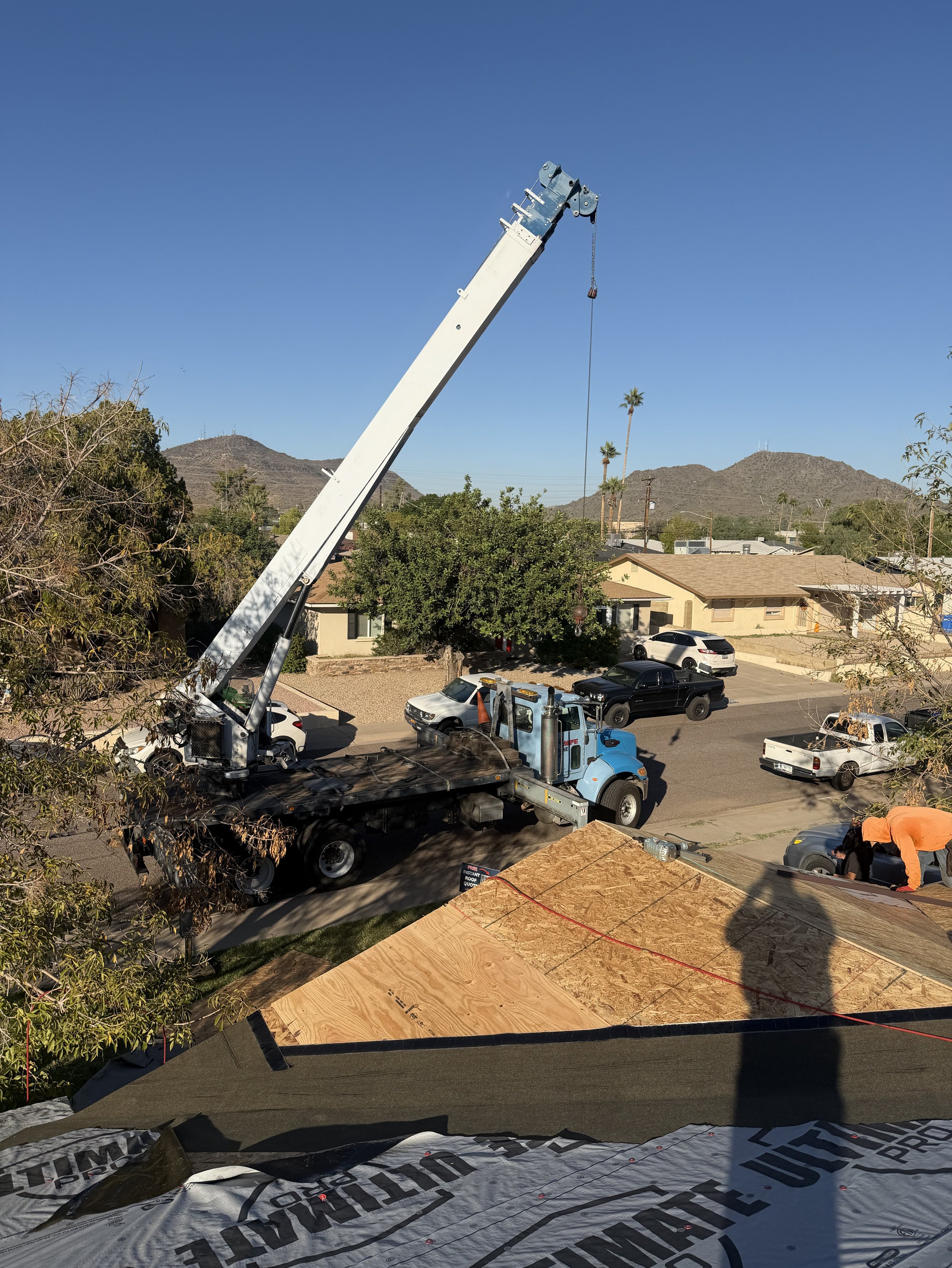 A blue crane truck operating on a residential street, lifting an object with a long boom arm. Construction workers are working on a roof with plywood sheathing. The background shows houses, trees, and distant mountains under a clear blue sky.