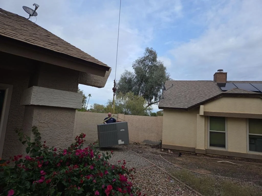 A man is seen hanging from a cable in a backyard, installing or repairing a large HVAC unit outside a house. The scene shows two houses, a flower bush in the foreground, and a cloudy sky overhead.
