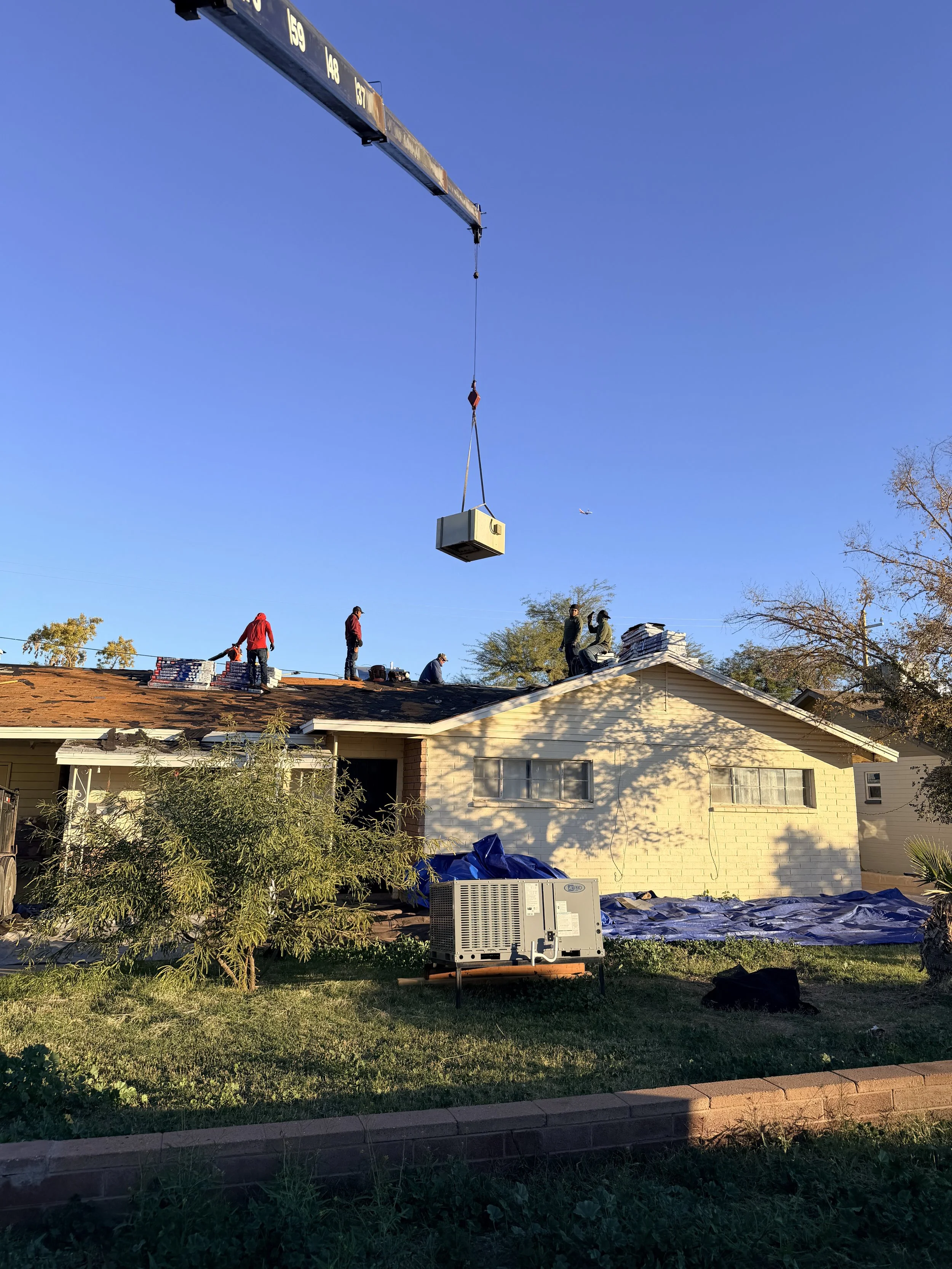 Construction workers on the roof of a house installing shingles, with a crane lifting a large box, and a clear blue sky in the background.