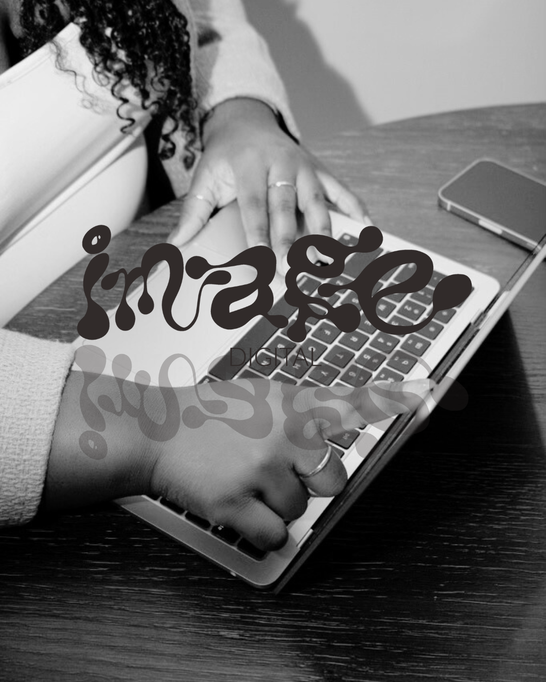 Person using a laptop with one hand on the keyboard and the other on an open notebook on a wooden desk, with a smartphone nearby.