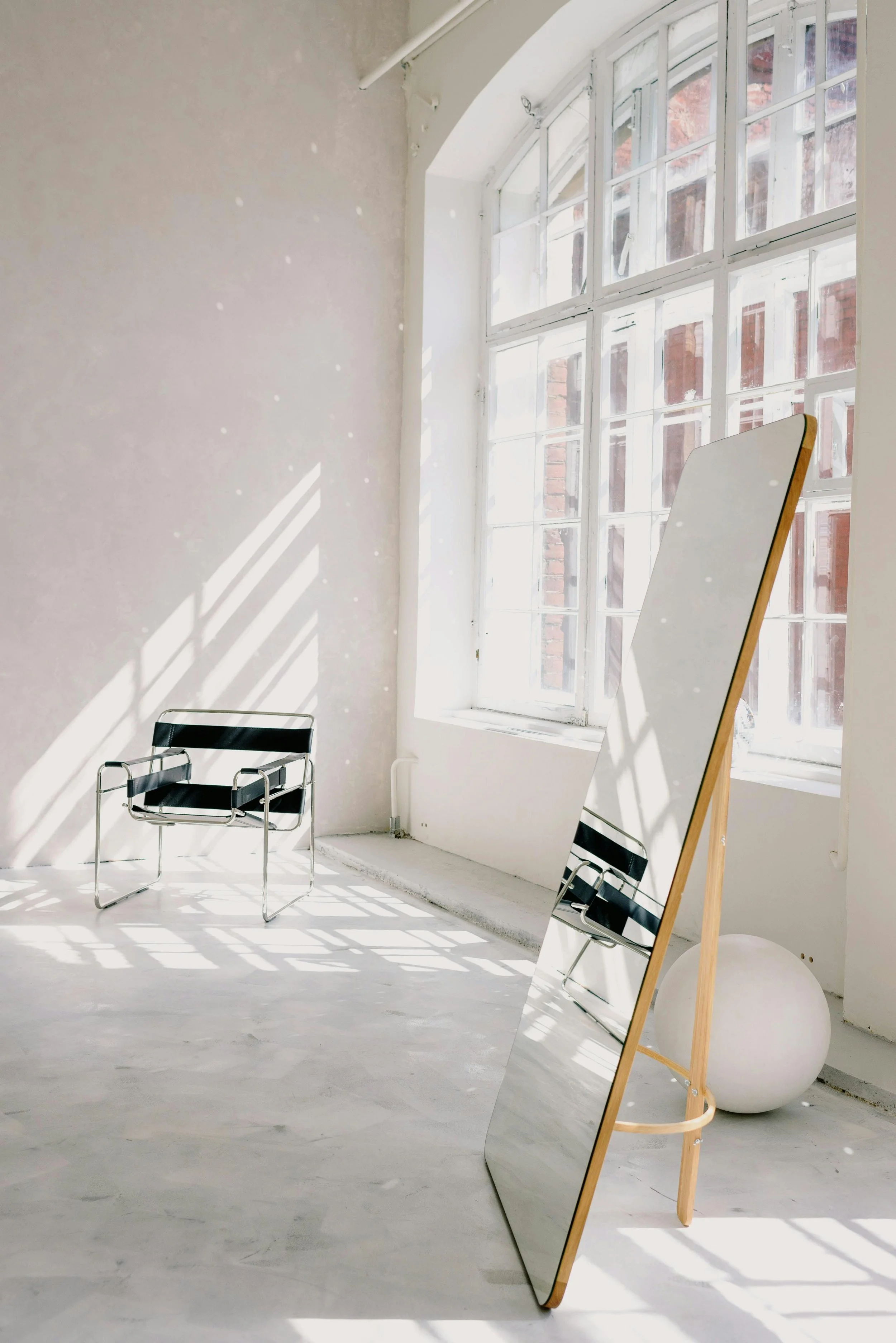 Minimalist interior with large window casting natural light and shadows, a full-length mirror on a wooden stand, a black chair with metal frame, and a white spherical object on the floor.