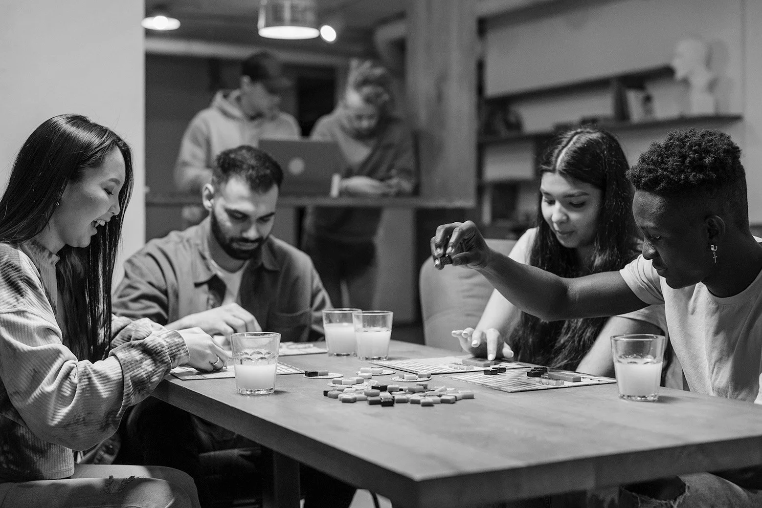 A group of five young people playing a board game at a table with glasses of drinks in a cozy indoor setting, with two people in the background working on laptops.
