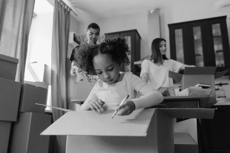 Young girl with curly hair coloring on a piece of paper inside a cardboard box, with two adults in the background surrounded by packing boxes.