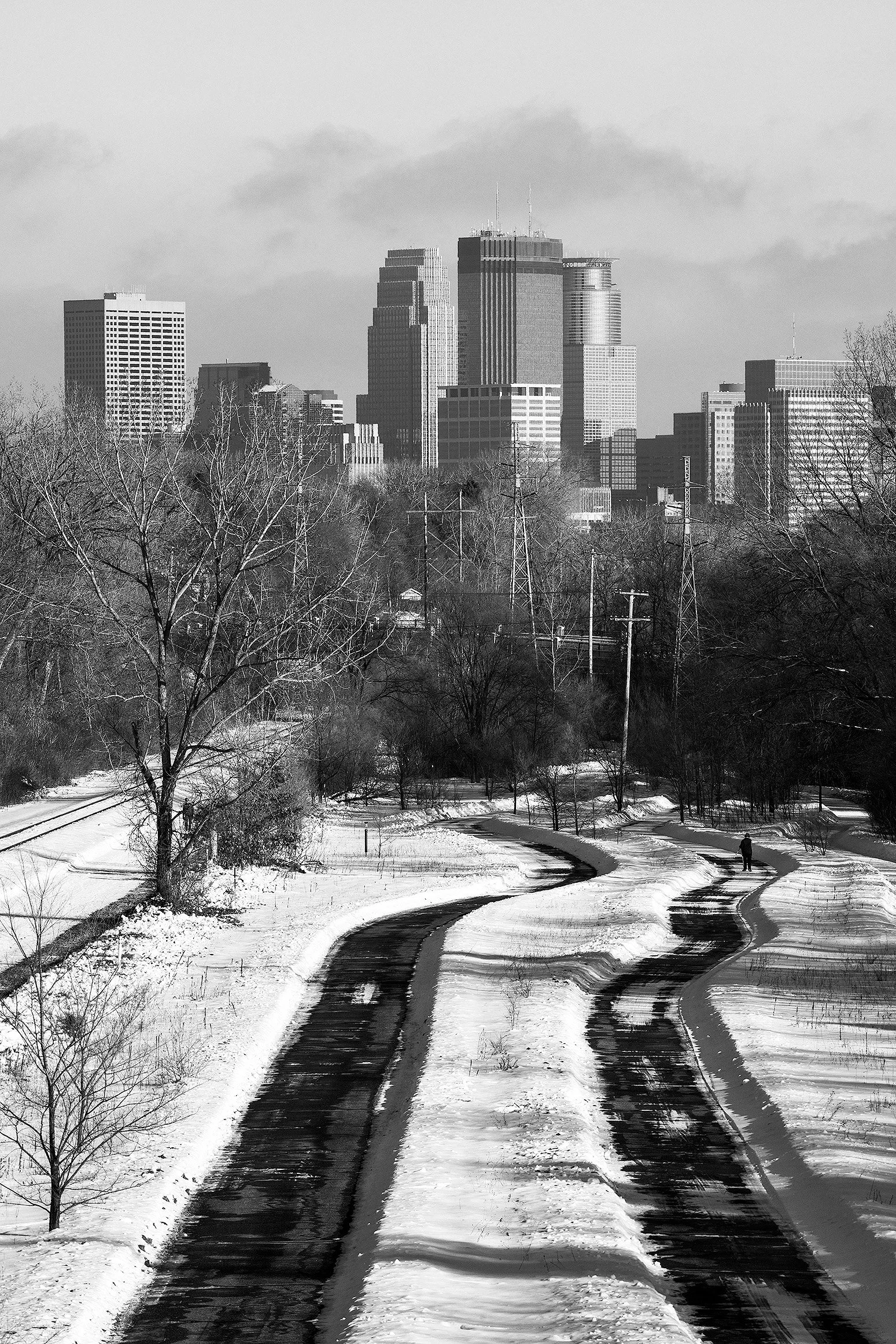 Black and white photo of a snowy park with winding pathways and a city skyline in the background.