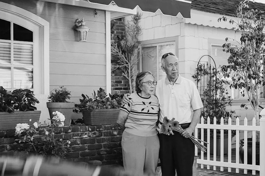 An elderly woman and man standing outside in a garden, with the woman holding a bouquet of flowers. They are in front of a house with plants and a white picket fence.