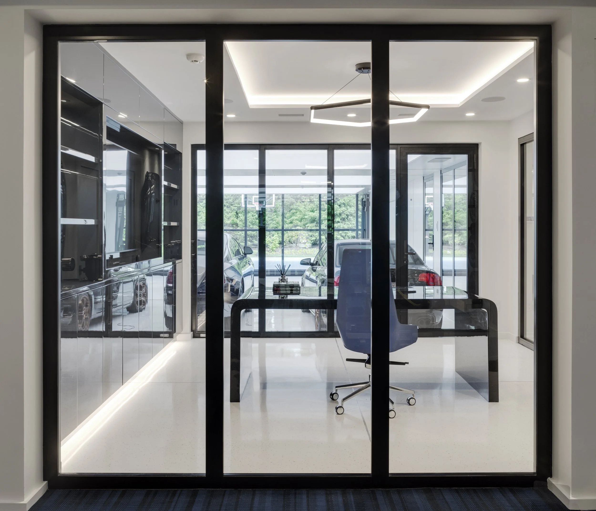 Modern home office with glass walls, black frame, blue chair, desk, window view of trees and cars, ceiling lights.