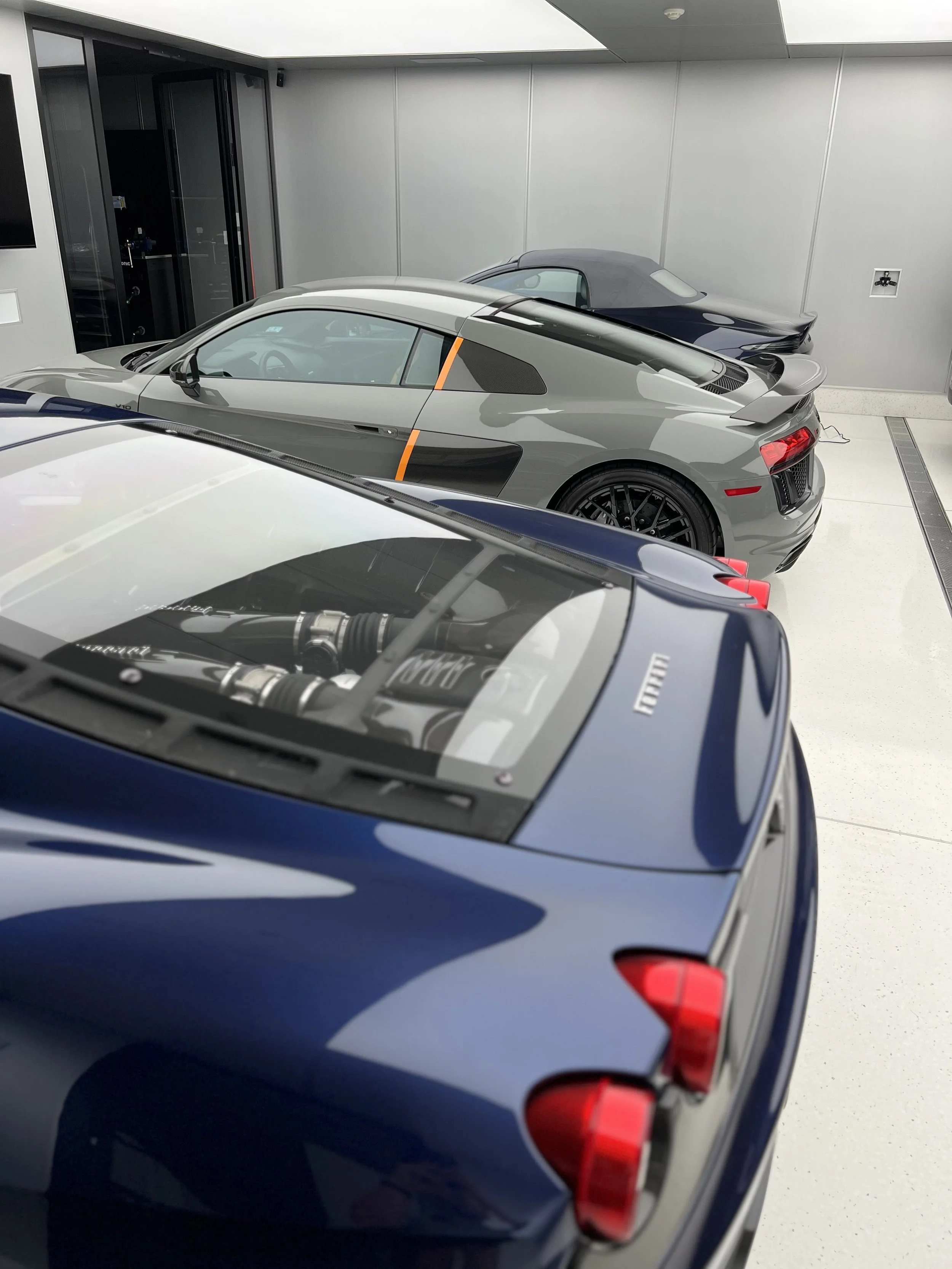 Three luxury sports cars parked indoors in a showroom or garage. The car in the foreground is blue, the middle car is gray, and the back car is dark blue or black.