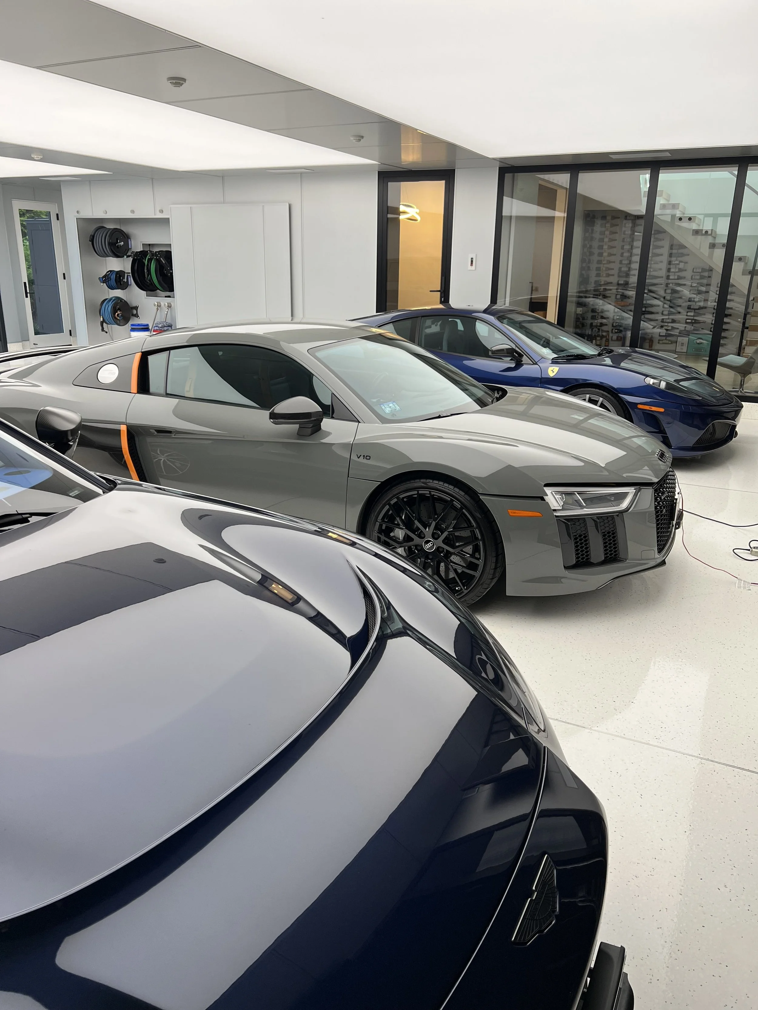 A garage with three luxury sports cars: a gray Audi R8 in the center, a blue Ferrari in the background, and a black Lamborghini in the foreground.