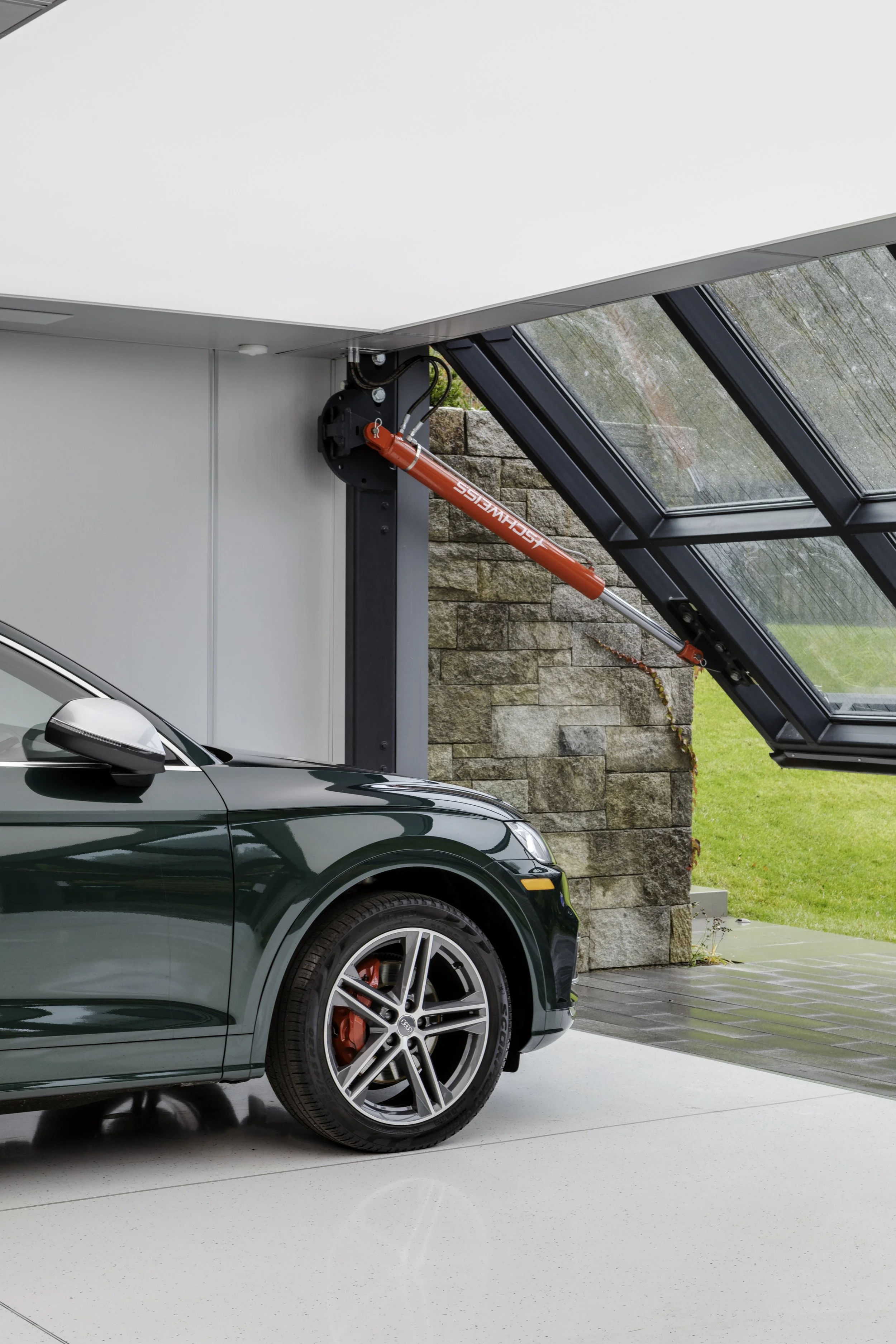 A green car parked inside a modern garage with a hydraulic lift raising the garage door. The door is made of glass panels and there is a stone wall outside.