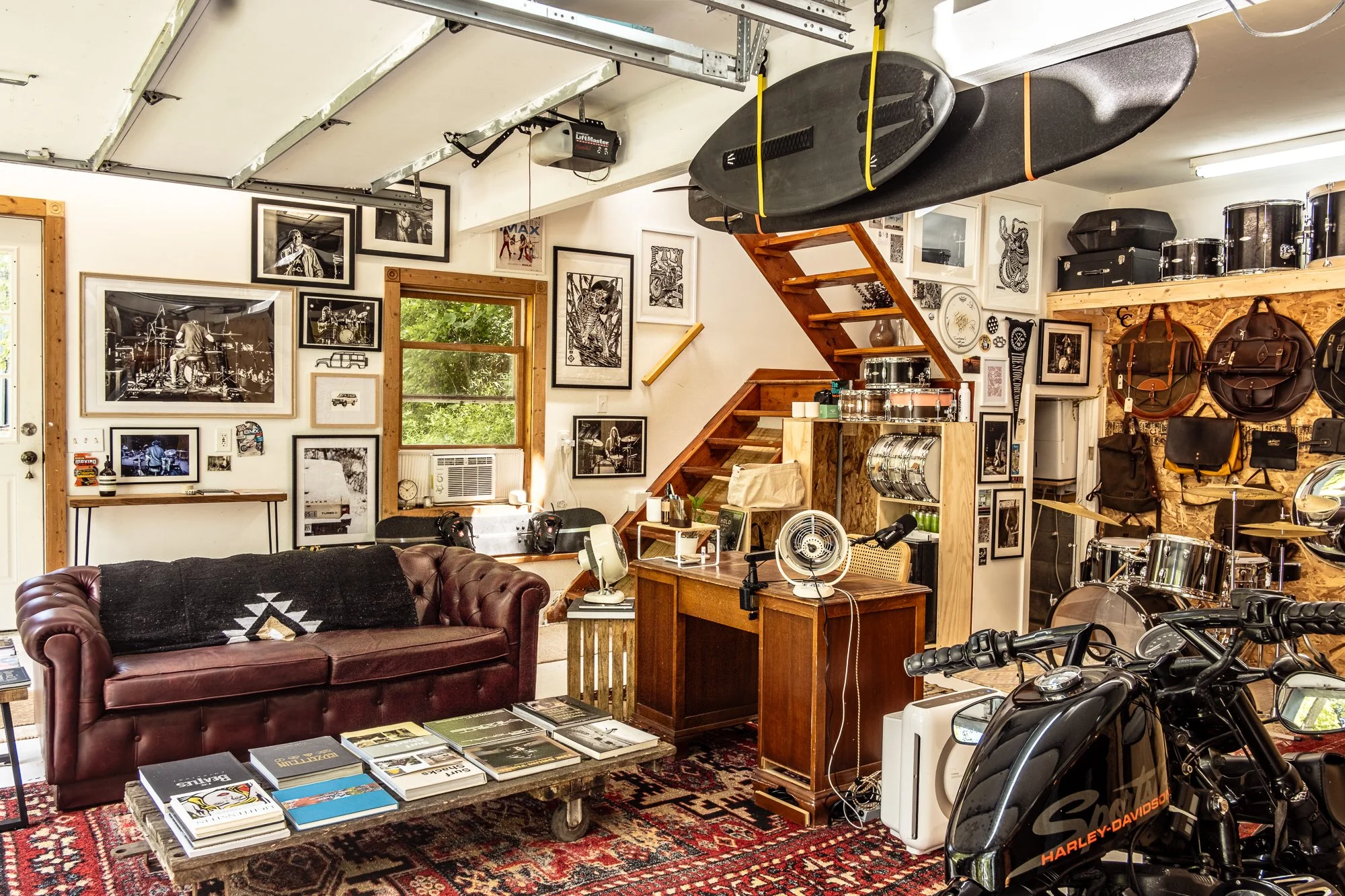 A room filled with musical instruments, artwork, and furniture, featuring black and white photographs on the walls, a leather couch, a coffee table with magazines, a motorcycle, and various musical equipment including drums, a surfboard, and audio ge