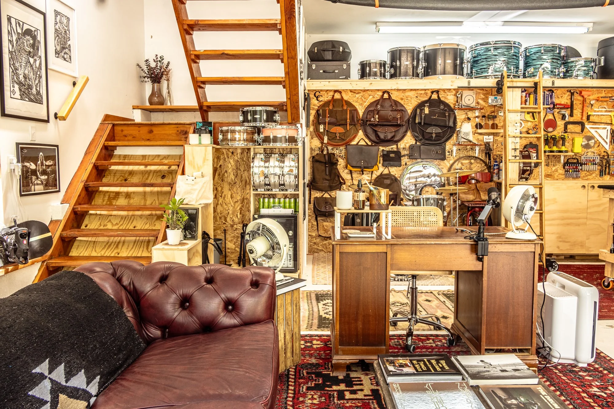 Interior of a creative workspace or studio with wooden stairs, wall-mounted backpacks and bags, shelves with drums, small instruments, and art supplies. There is a wooden desk with tools, a vintage leather sofa, and books on a patterned rug. The spac