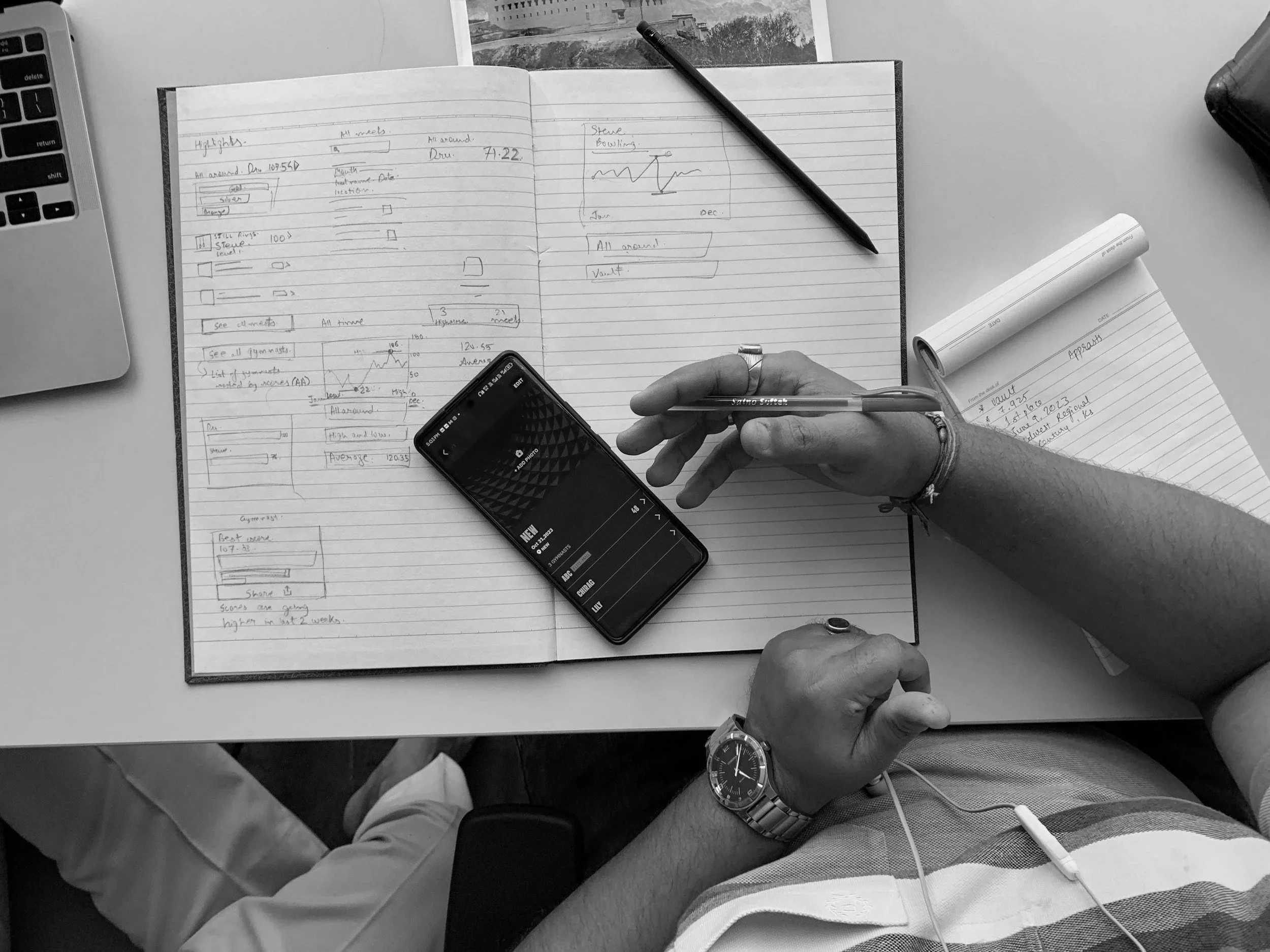 A person with a watch on their left wrist using a pen to write in a notebook. The notebook contains notes, diagrams, and graphs. A smartphone, a laptop, and a rolled-up notepad are also on the desk. Another person is partially visible sitting nearby.